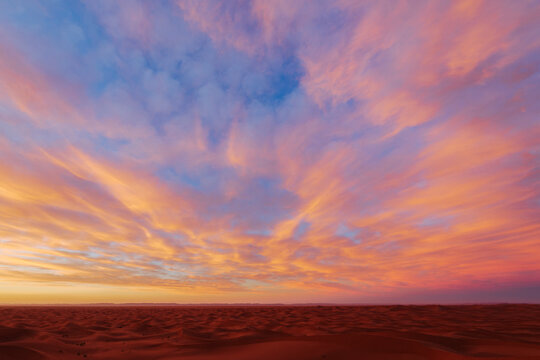 The sky at sunrise above the Chaggaga Sand Dunes, light clouds reflecting red and orange light against blue sky.