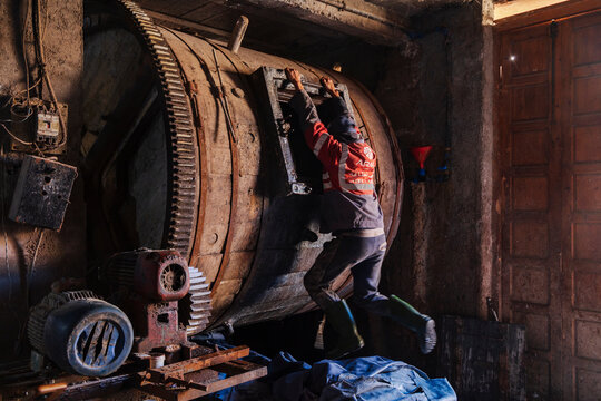 A man turning a large wooden cask or barrel by pulling on a ledge, cogs and wheels and old metalwork in a tannery.