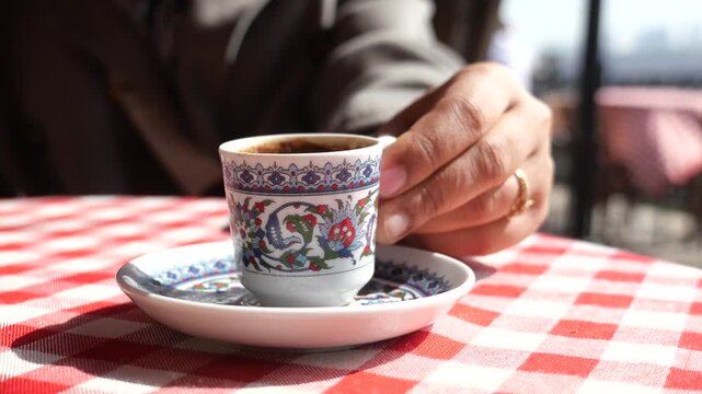 A man's hand gracefully lifts a small, intricately patterned Turkish coffee cup from its matching saucer on a red and white checkered tablecloth