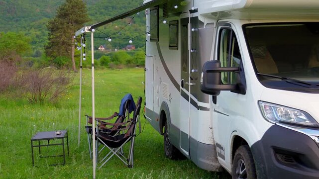 Modern motorhome standing in a scenic green valley with a camping chair and table set up under the awning, showcasing a peaceful van life concept amidst nature on a beautiful spring day