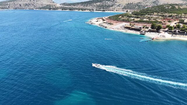 Drone shot of motorboats leaving white foam trails in clear deep blue water. Scenic summer seascape with green coastline, sandy beach, and resort infrastructure against mountains background.