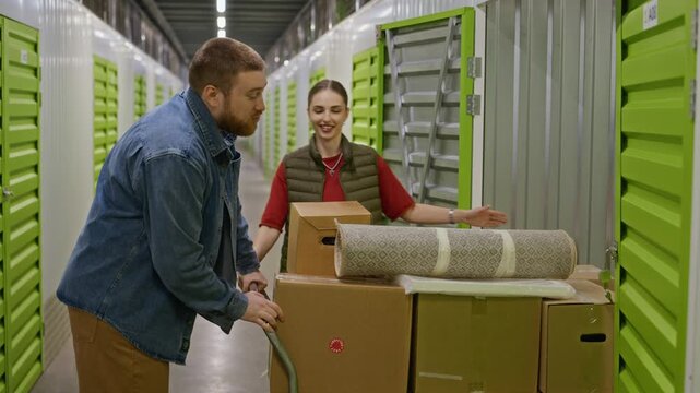 Young adult Caucasian couple pushing cart with boxes and carpet into self storage unit. Perfect for promoting relocation services, moving companies, and personal storage solutions