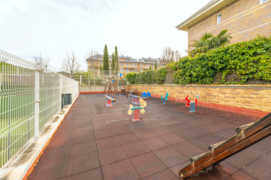 Community children's playground with spring rockers and rubber flooring, enclosed by fencing beside a green hedge and brick building.