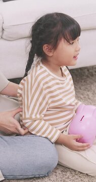 Close up of a cute Asian girl holding a pink piggy bank while mother hugs her lovingly at home.