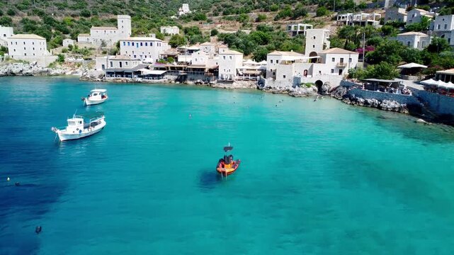 Drone footage of traditional fishing boats floating in the crystal-clear turquoise waters of Limeni Bay, Mani, Greece, with historic stone buildings along the Mediterranean coastline.