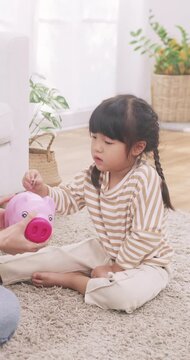 Asian cute little girl saving money by putting coins into a pink piggy bank with her mother at home.
