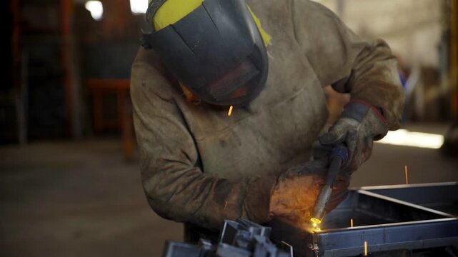 A cinematic close-up of a professional welder wearing a protective mask, leaning over to weld a complex metal framework, creating bright sparks inside a dark factory setting.