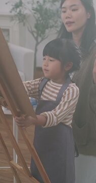 Asian mother teaching her young daughter how to paint on an easel at home.