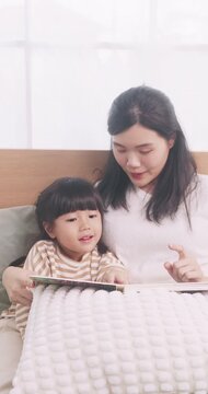 Asian mother sitting on bed reading a storybook with her cute young daughter at home.