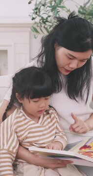 Asian mother sitting on bed reading a storybook with her cute young daughter at home.