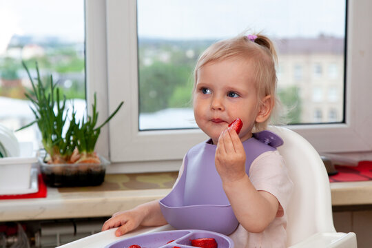 A young child sits at a table and enjoys fresh strawberries. Family members are present and smiling. The sun is shining through the window, creating a warm atmosphere.
