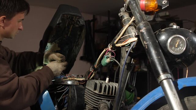 Man in work gloves carefully connecting intricate electrical wires on a disassembled vintage motorcycle, performing maintenance and repair in a workshop setting