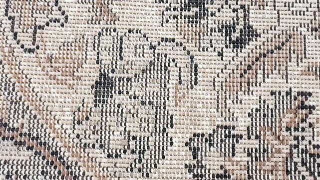 Detailed close-up of an aged woven carpet featuring a traditional geometric pattern in beige, cream, black, and brown tones. The camera slowly moves across the textured surface