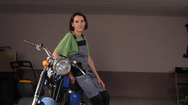 Young woman wearing work overalls and green t shirt sitting confidently on her vintage blue motorcycle inside a garage, representing female empowerment in a traditionally male profession