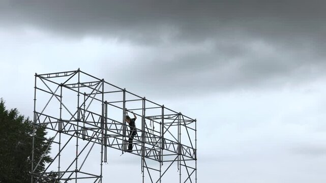 Construction worker securing ropes on tall metal scaffolding under heavy overcast sky, standing on horizontal brace with safety harness and helmet in industrial outdoor setting