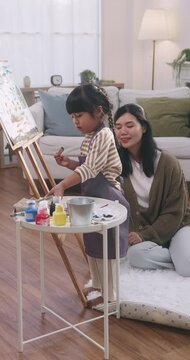 Asian mother teaching her young daughter how to paint on an easel at home.