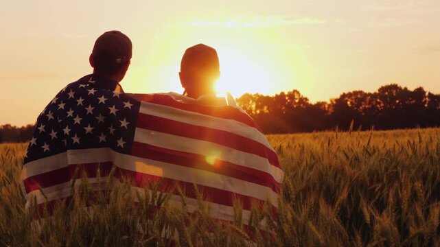 Two people silhouette covered with united states flag sitting in wheat field at sunset.