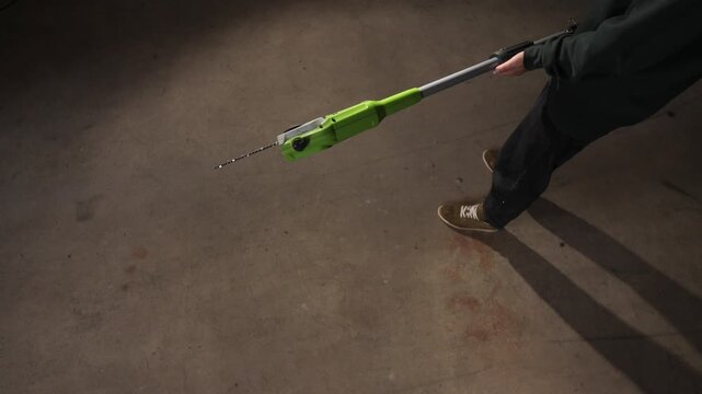 High angle view person is holding a long reach electric pole saw. Featuring a bright green main body and a chain blade. While standing on a concrete ground