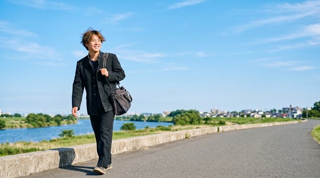 青空の下、堤防を笑顔で歩く学ラン姿の男子高校生