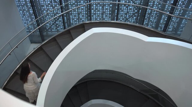 Overhead modern spiral staircase with woman descending along curved white balustrade beneath patterned blue glazing in minimalist interior, with staircase, spiral, balustrade, handrail detail