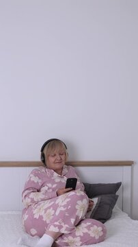 Senior Woman Listening to Music in Bed. Senior woman in pajamas sitting in bed at home and listening to music on smartphone with headphones.