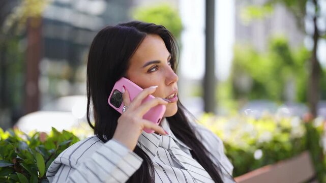 A woman is sitting on a bench in a city park. She is talking on her pink phone, surrounded by green plants and trees. The sun is shining on a clear day.