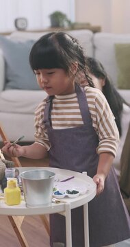 Asian mother teaching her young daughter how to paint on an easel at home.