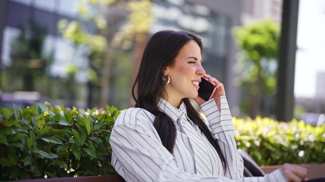 A woman is sitting on a bench in a park. She is wearing a striped shirt and is using her phone to talk. The park has trees and buildings in the background.