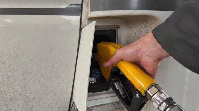 Close-up view of a man's hand inserting a yellow diesel fuel nozzle into the tank of a dirty white vehicle at a gas station, highlighting the rising cost of fuel and transportation