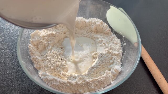 Close-up of pouring milk into a glass bowl with flour to make dough. baking bread, pizza, or pastry dough in a home kitchen.