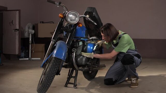 Young female mechanic crouches, intently focusing and illuminating the engine of a classic blue motorcycle with a flashlight, performing maintenance in a home garage