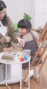 Asian mother teaching her young daughter how to paint on an easel at home.