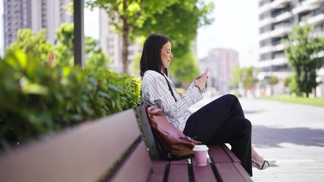 A woman sits on a bench in a city park. She is looking at her phone. There are tall buildings and trees around her. A coffee cup is placed beside her on the bench.