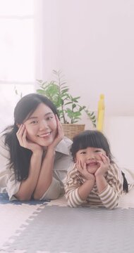 Happy Asian mother and daughter lying on floor resting chin on hands smiling at camera.