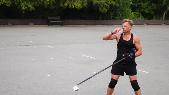 A middle-aged man drinks water after a roller hockey practice on an asphalt rink. He quenches his thirst and feels satisfied after a well-executed workout. Slow motion 4k footage.