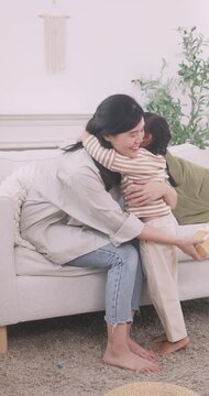 Little Asian girl giving gift box to mother and hugging her on sofa.