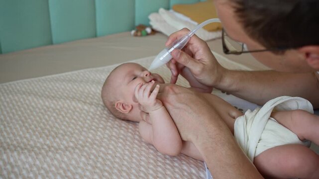Attentive father providing gentle care for his newborn baby, using a mechanical nasal aspirator to clean the infant's stuffy nose and relieve congestion for easier breathing at home