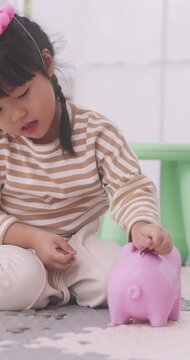 Little Asian girl putting coins into pink piggy bank on floor at home.