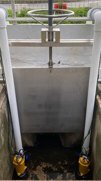 Close-up of a stainless steel manual sluice gate valve installed in a wastewater treatment facility. Industrial flow control equipment with handwheel mechanism used for regulating water level, drainag