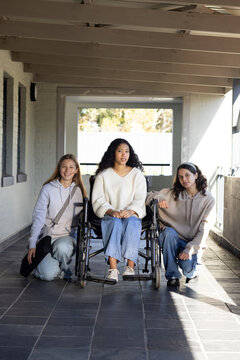 Diverse teenage female friends kneeling and posing around manual wheelchair on tiled porch