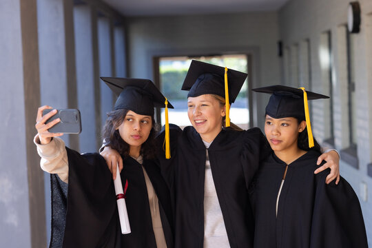Diverse female graduates taking selfie with phone in corridor, wearing gowns, caps, holding diploma