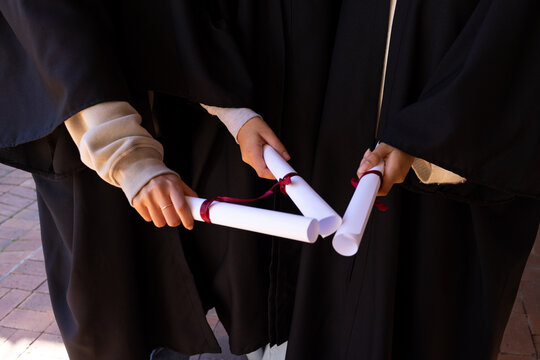 Female grads teen child holding 3 white diplomas wearing black gowns visible sleeves on brick path