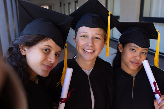 Three teenage female students posing in corridor with black gowns, caps, yellow tassels, diplomas
