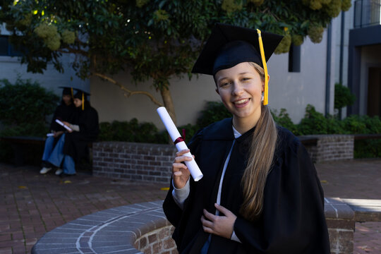 Youth female grad standing, holding diploma, wearing cap with gold tassel and gown in campus quad