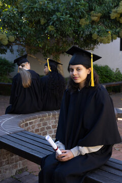 Diverse teenage female grads sitting on bench in quad, holding red-ribbon diploma, wearing caps