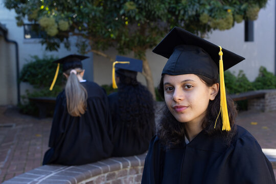 Diverse female grads and child sitting on campus in gowns, caps with yellow tassels, copy space
