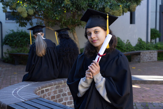 Diverse teenage graduates wearing black gowns caps posing on campus holding red-ribboned diploma