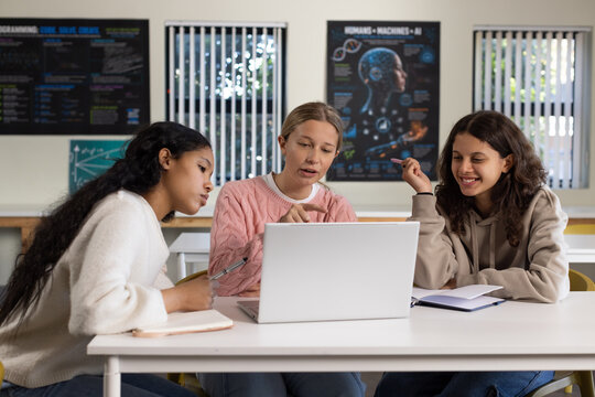 Female teacher guiding female teenage students at school, pointing at laptop and writing notes