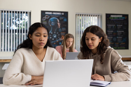 Teen female students wearing cream sweater beige hoodie leaning at school studying on silver laptop