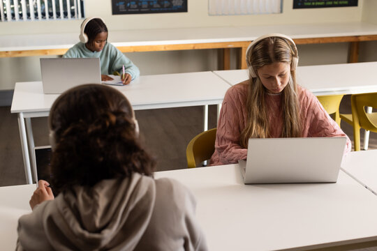 Diverse students using laptops and headphones in classroom, pink-sweater teen right, copy space